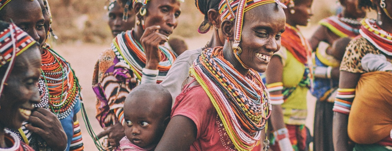 A group of people with dark skin tones all wearing brightly colored clothing, multiple beaded necklaces, and beaded caps