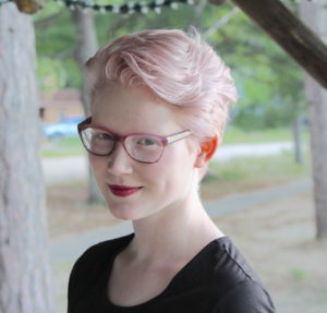 Val has light pink hair, wears glasses, and is smiling. They looking slightly to the left to look at the camera with trees in the background.